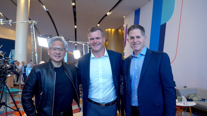 Three men pose together at what appears to be a corporate or industry event, with one in a black leather jacket on the left, one in a light blue shirt in the center, and another in a blue suit jacket on the right. The professional setting with lighting equipment in the background suggests this could be a media interview or conference, potentially relating to $IREN, an AI/data center company focused on GPU computing.
