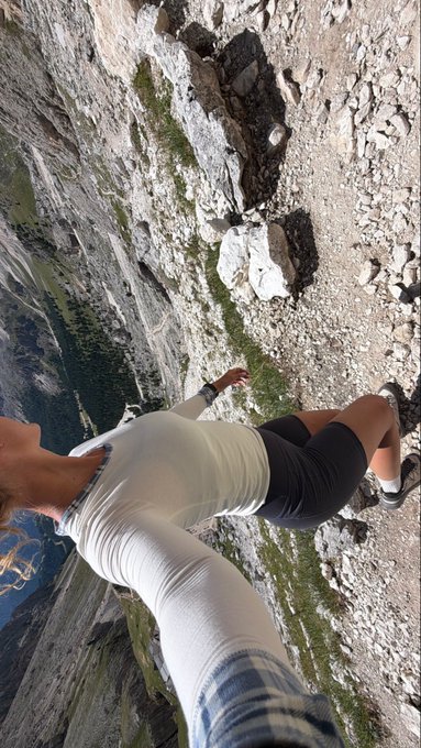 A selfie taken at a dizzying angle on a steep rocky cliff, showing the person in a white long-sleeve shirt and black shorts with a forested valley far below.