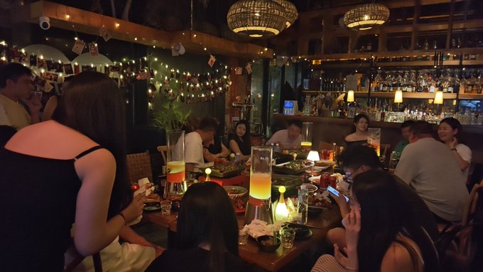 A dimly lit, cozy bar is packed with patrons sitting at a long wooden table, illuminated by warm string lights, decorative lanterns, and a disco ball.