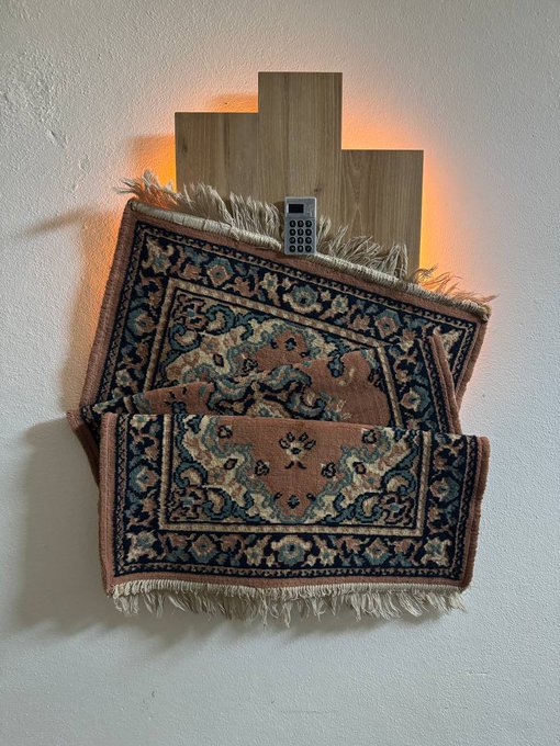 Two small Persian-style rugs with intricate pink and navy patterns and fringed edges are mounted on a wall against a wooden Bitcoin-shaped backing, with a Coldcard hardware wallet displayed at the center—visually representing the artist's "Coldcard Rug Pull" concept by fusing cryptocurrency hardware security with traditional textile art.