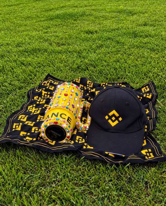A gold "BNCE" mug embellished with colorful rhinestones sits next to a black cap featuring the Binance logo, both resting on a matching black and yellow patterned cloth spread across green grass—perfectly capturing the post's "Adding some sparkle to our Sunday" vibe with its bedazzled Binance-branded accessories.