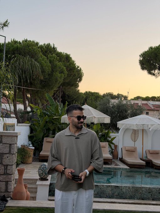 A man wearing sunglasses and an oversized olive green shirt stands in a resort area at sunset, with a pool, lounge chairs, and decorative umbrellas behind him.