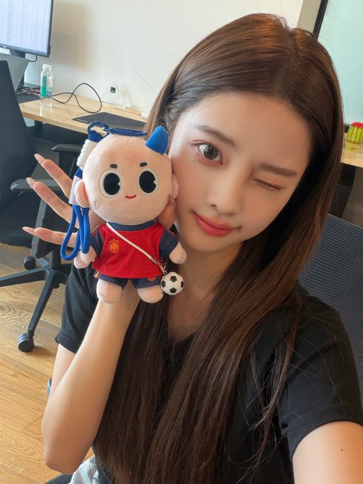 A selfie of a young woman winking at the camera while holding a small plush toy of a baby character in a red soccer uniform with a soccer ball charm, in what appears to be an office setting.