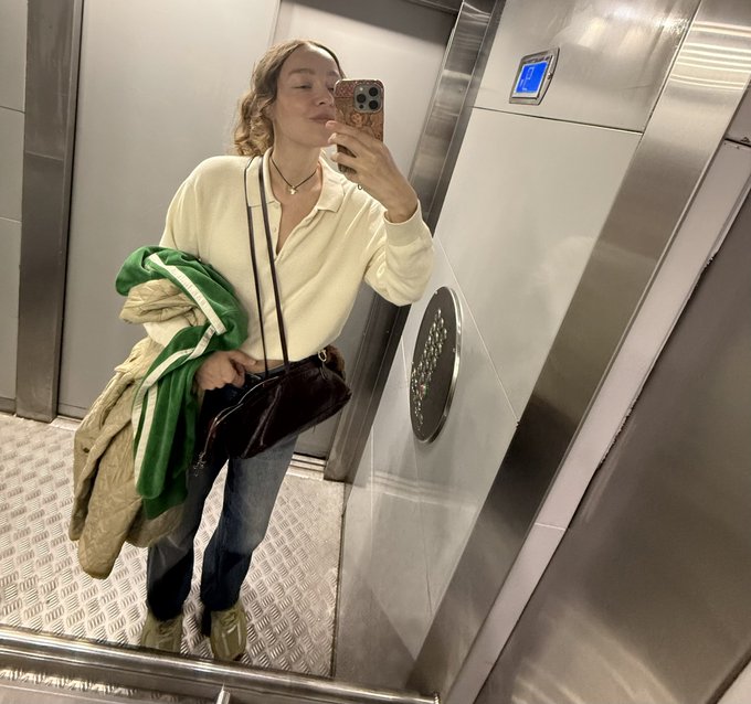 A mirror selfie in an elevator of a woman wearing a cream zip-up top and jeans, holding a green and white jacket and a brown crossbody bag.