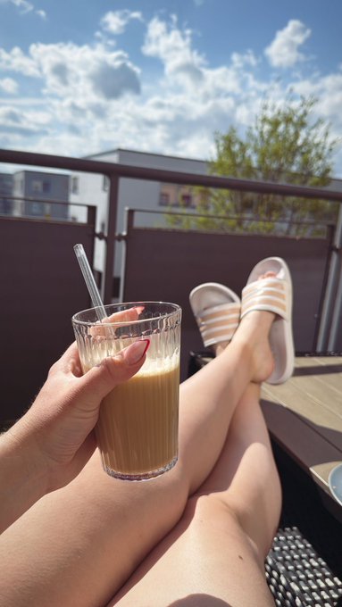A first-person view of someone relaxing on a sunny rooftop or balcony, holding an iced coffee drink in one hand while wearing white Adidas slide sandals, with their legs crossed.