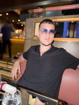 A man wearing sunglasses and a black polo shirt sits in a booth at what appears to be a restaurant or bar.