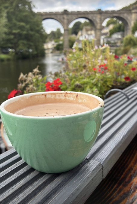 A green ceramic cup filled with a frothy beverage sits on a metal railing, with red flowers and an old stone railway viaduct visible across a river in the blurred background.