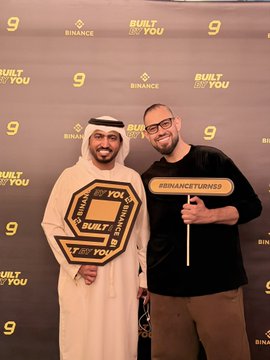Two men pose in front of a "Built for You" and "Binance 9" branded step-and-repeat backdrop, with one holding a "#BINANCETURNS9" sign. The image captures the celebratory 9th anniversary event referenced in the caption, highlighting Binance's milestone of nine years of innovation.