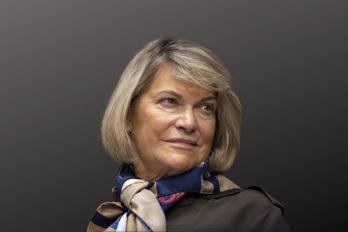 A woman with shoulder-length blonde hair wearing a patterned silk scarf and dark jacket poses against a gray background.