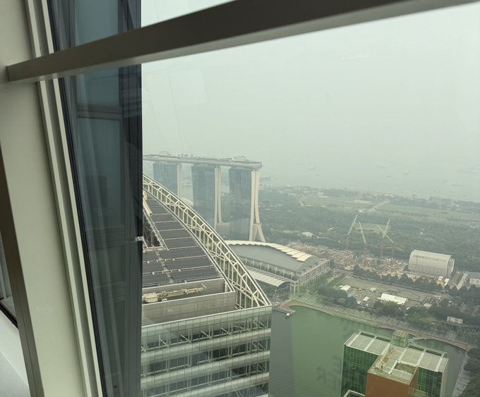 A hazy view from a high-rise window reveals the iconic Marina Bay Sands hotel with its distinctive SkyPark and rooftop infinity pool, alongside the surrounding Singapore skyline and harbor. The image likely relates to the accompanying post by suggesting a meeting location or the upcoming Near Protocol team gathering in Singapore.