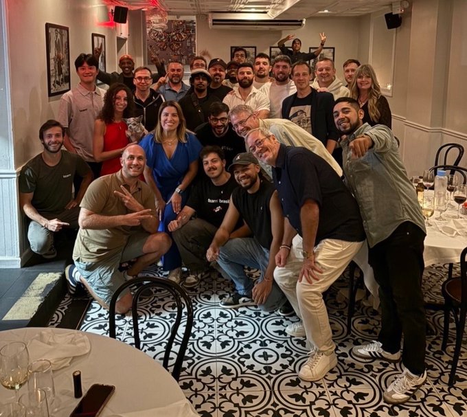 A large group of approximately twenty people poses for a group photo at a gathering in a restaurant with patterned tile flooring. Some individuals are making peace signs, and a small dog is being held in the crowd.