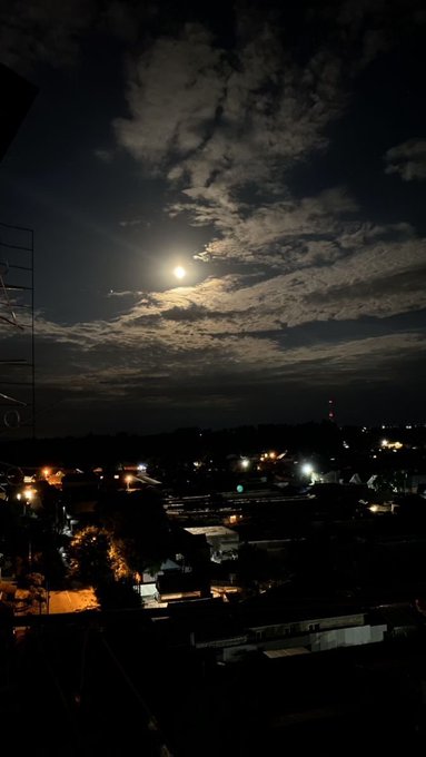 The image shows a view of an urban city at night with a full moon shining brightly behind it, creating a beautiful scene against the dark sky. The streetlights cast their glow onto the buildings, making them appear illuminated. A few cars can be seen parked along the road in front of the buildings, and there is a tall tower in the distance.
