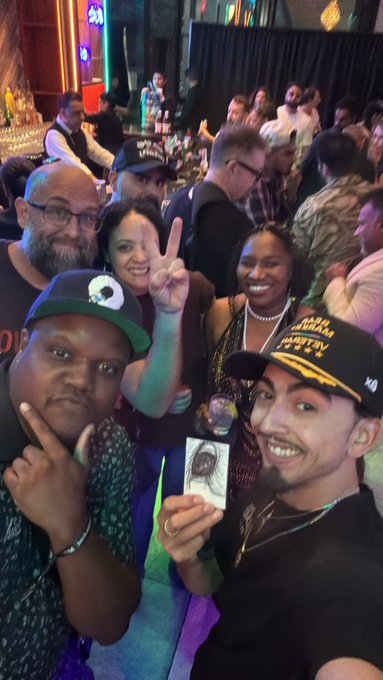 A group selfie of people at a crowded event, with one person wearing a 'Starship Troopers' cap and another holding a sketch drawing.