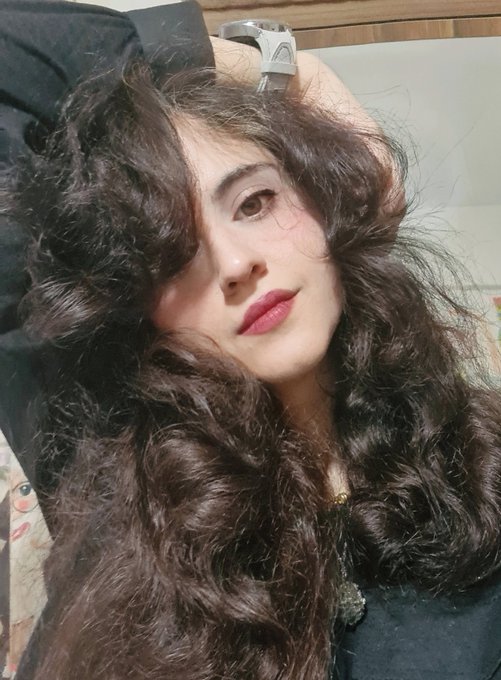 A young woman with long, voluminous curly dark hair and bold makeup rests her arm behind her head, wearing a watch, in a casual selfie that serves as her personal introduction as the owner of the account.