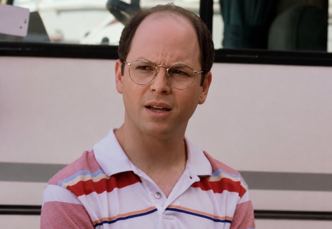 A man wearing wire-rimmed glasses and a striped polo shirt standing in front of what appears to be a bus, recognizable as the character George Costanza from Seinfeld.