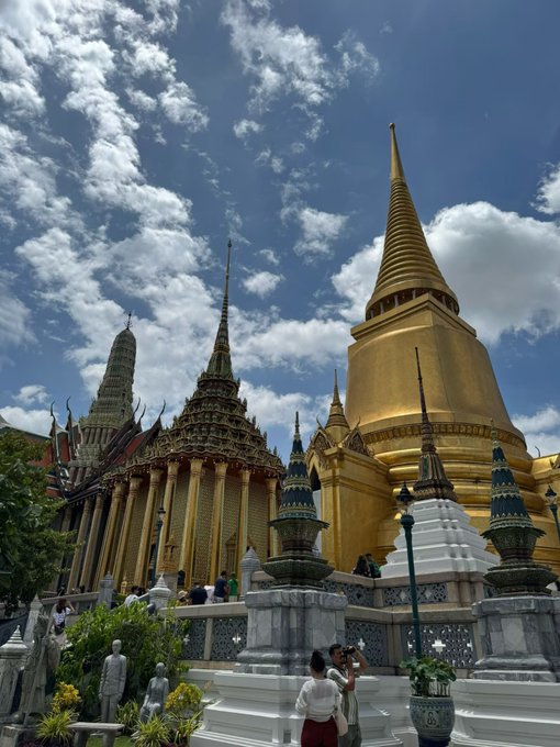 A low-angle photograph captures the Grand Palace in Bangkok, featuring a large golden chedi and ornate spires against a partly cloudy blue sky, with tourists and statues visible at the base.