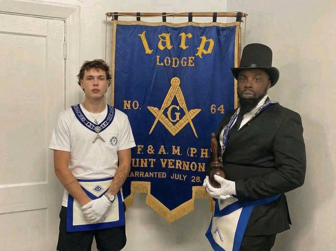 Two men stand in front of a blue Masonic banner reading "Harp Lodge NO. 64 F.&A.M. PH. Mount Vernon", both wearing ceremonial aprons and white gloves, with the man on the right in a top hat.