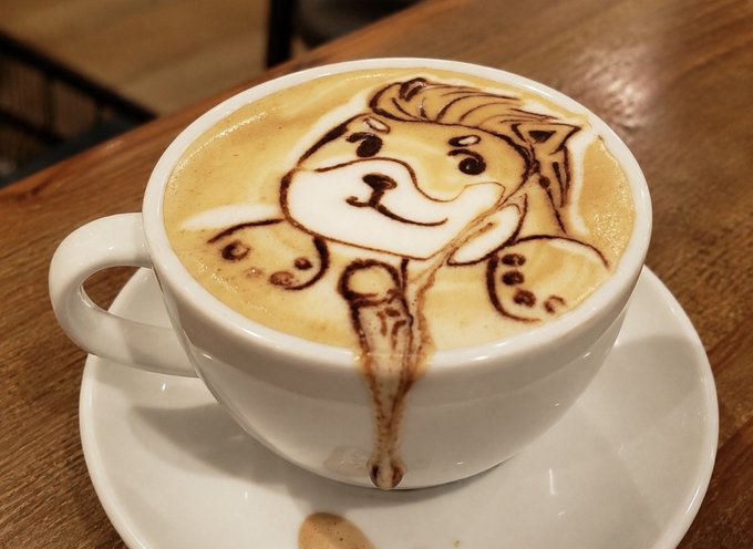 A close-up of a cappuccino with intricate latte art depicting a cartoon dog's face created with chocolate syrup, served in a white cup on a saucer atop a wooden table.