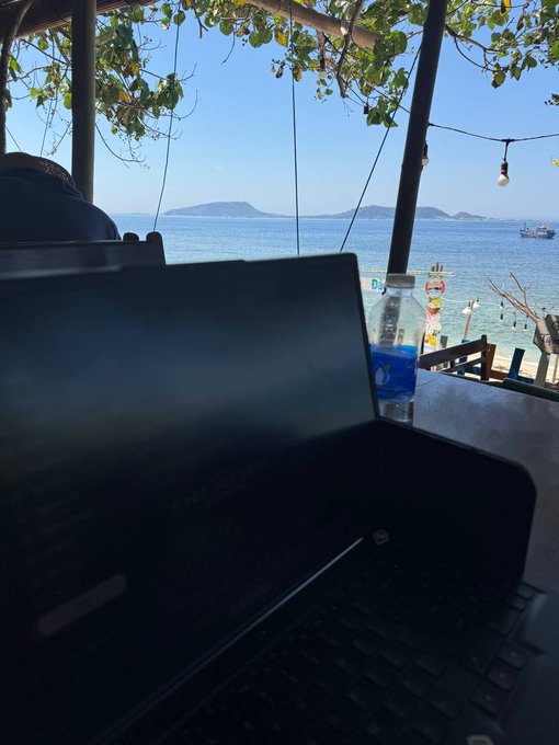 iced tea and a glass of water accompany an open laptop on a table, overlooking the ocean with a clear blue sky and palm trees in the background.