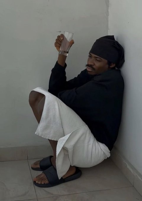 A man wearing a black head wrap and shirt sits slumped against a wall, holding up an empty glass in a gesture of despair or frustration. The image humorously illustrates the post's lament about financial hardship, suggesting the empty glass symbolizes the lost opportunity of not investing in Bitcoin.