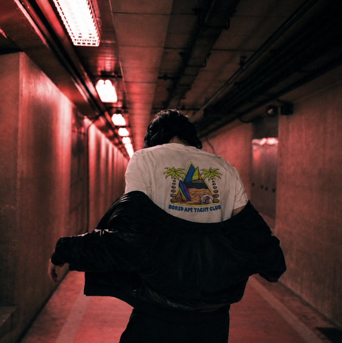 A person walks down a red-lit industrial tunnel, showcasing a white t-shirt reading "BORED APE YACHT CLUB" with its iconic blue ape logo and palm trees. The image's moody, atmospheric setting complements the "Generational Run" theme, evoking a sense of legacy and movement through a bold, stylized environment.