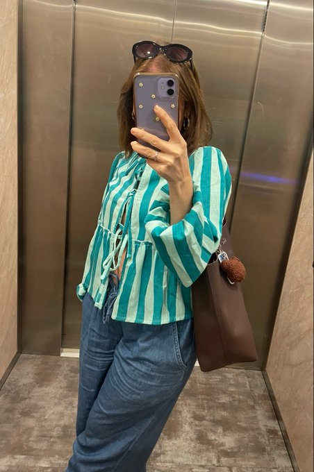 A mirror selfie taken in an elevator showing a person wearing a teal and white striped blouse, blue jeans, sunglasses on their head, and carrying a brown tote bag with a teddy bear charm.