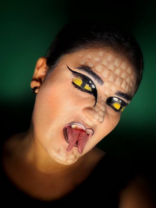 A close-up portrait of a woman with snake/reptile-inspired makeup, including scale patterns on her forehead and cheeks, striking yellow eyeshadow, and a forked tongue sticking out against a green background.