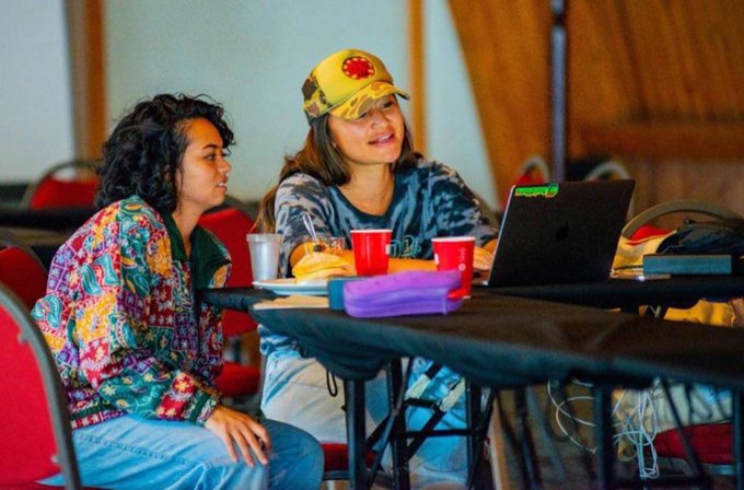 Two women sitting at a table looking toward a laptop screen, one wearing a colorful patterned jacket and the other in a camouflage shirt and yellow floral cap, with red plastic cups and a purple pencil case on the table.