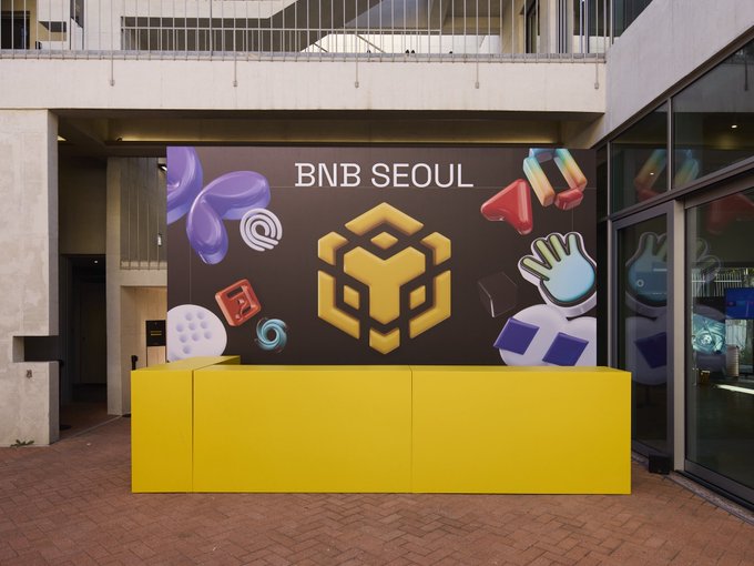 A large BNB Seoul promotional display featuring the BNB Chain logo and colorful 3D icons of various crypto-related objects sits behind a bright yellow counter in an empty outdoor venue, capturing the quiet setup before the event begins.