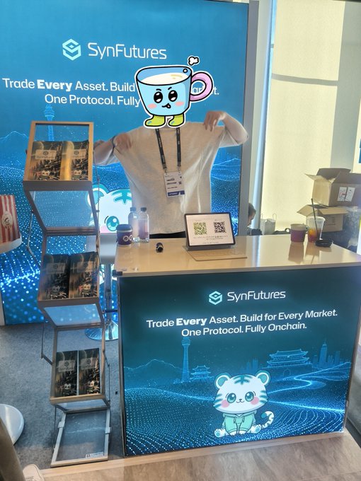 A person poses with a peace sign behind a SynFutures booth counter at KBW2026, with the brand's slogan "Trade Every Asset. Build for Every Market. One Protocol. Fully Onchain." displayed on the back wall and a digital screen featuring a cartoon tiger mascot. A cute cartoon cup character has been edited over the person's face, matching the post's playful tone celebrating the SynFuturesDefi booth experience.