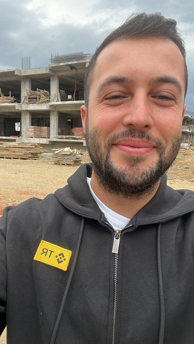 A selfie of a smiling bearded man wearing a black hoodie with a yellow patch, standing in front of an unfinished building construction site.
