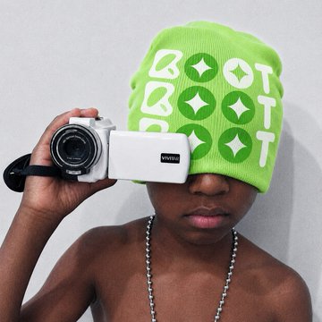 A person wearing a bright green BOTT BOTT beanie holding a white compact camera up to their face.