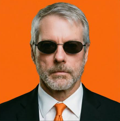A portrait of an older man with gray hair, a gray beard, and dark sunglasses, wearing a black suit, white shirt, and orange tie against a bright orange background.