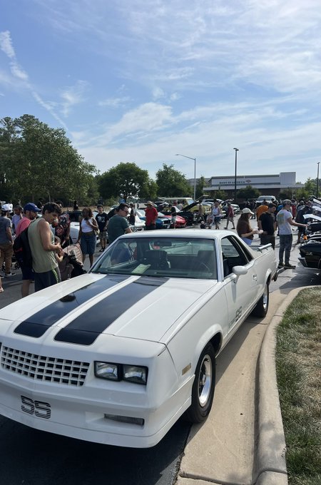 Outdoor photograph of a white Chevrolet El Camino SS with black racing stripes parked at what appears to be a car show, with crowds and other vehicles visible in the background.