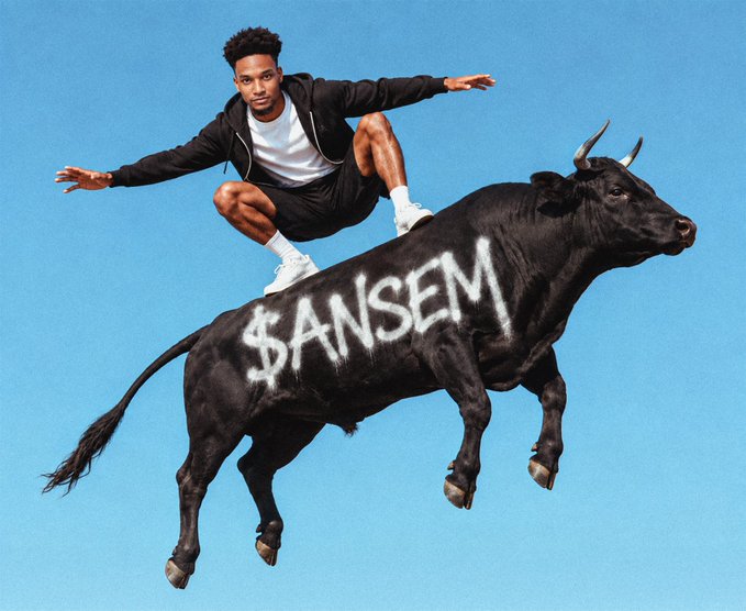 A young man in athletic wear crouches on the back of a leaping black bull with '$ANSEM' written on its side against a blue sky.