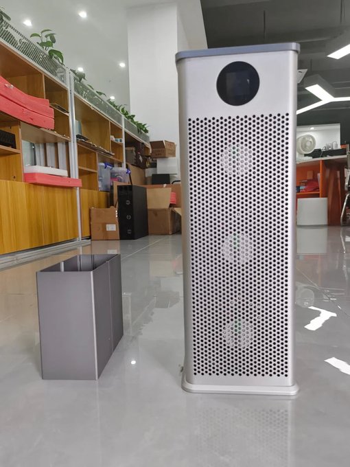 A tall, silver air purifier with a perforated front grille and a circular display sits on a tiled floor in what appears to be a showroom or office, with another unit lying on its side nearby.