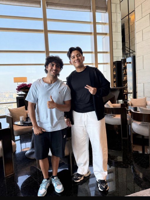 Two smiling young men posing indoors in a high-rise lounge area with large windows; one wears a white t-shirt and shorts while the other wears a black sweater and white pants.