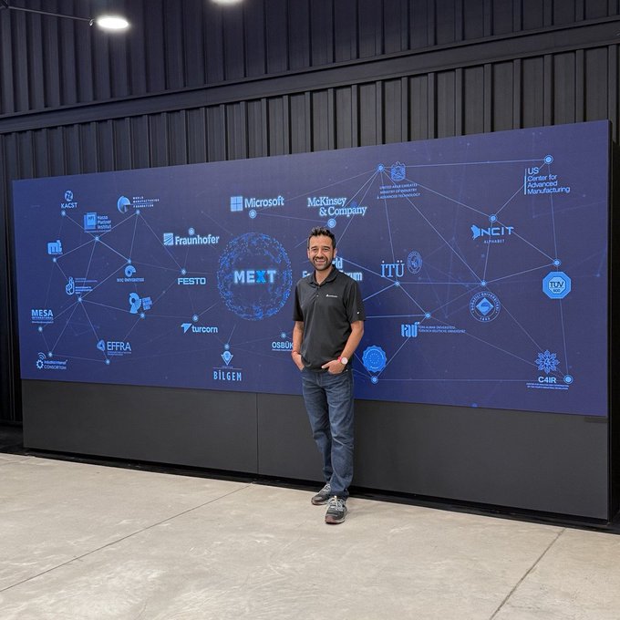 A man in a black polo shirt stands smiling in front of a large blue digital display showing various company and university logos networked together, including Microsoft, McKinsey & Company, Fraunhofer, and ITU.