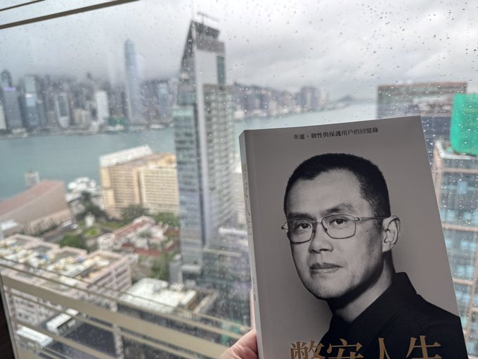 A hand holds a book featuring a man's portrait with Chinese characters and a view of a rainy cityscape through a window in the background.