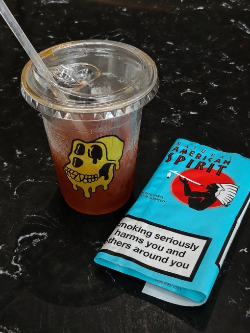 The image features a cup of tea with a skull design, placed next to an ashtray filled with cigarette butts. The cup has a straw sticking out of it, while the ashtray contains numerous cigarette butts. The background is black, which contrasts with the yellow and blue colors on the ashtray. The post includes text that reads "GM BAYC FAM 下面做持乐蓝V互关 今天的你喝啡了吗 请在评论里分享出来".