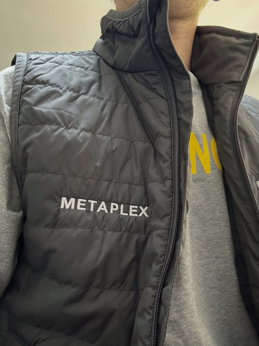A person models a black puffy vest with 'METAPLEX' printed in white letters across the chest, layered over a gray sweatshirt.