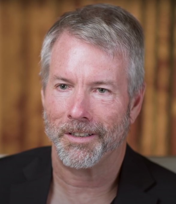 A close-up portrait photograph of a gray-haired, bearded man in a dark suit jacket against a blurred background.