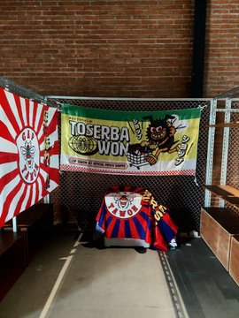 A colorful merch display for “TOSERBA WON” is set up on a brick wall, with a red-and-white striped shirt, a multicolor jacket, and a large banner featuring the brand name and graphics. It relates to the post’s “New merch drop” announcement as the pictured items appear to be the new merchandise being promoted.