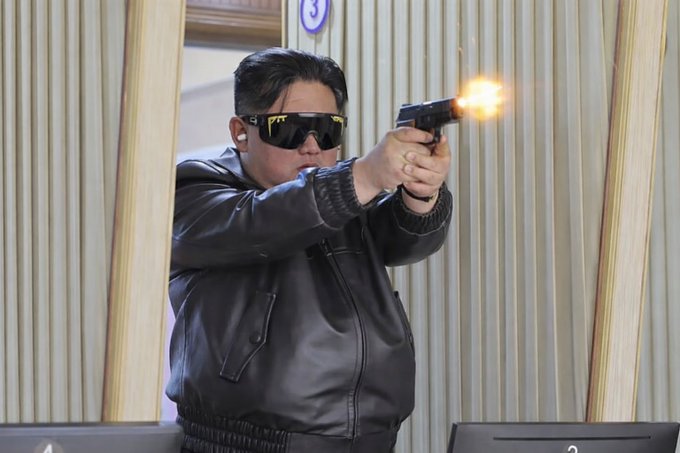 A man wearing sunglasses and a black leather jacket fires a handgun in an indoor shooting range, with a muzzle flash visible from the barrel.