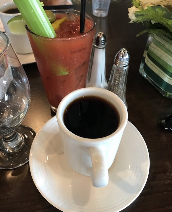 A close-up of drinks on a dark table: a white cup of black coffee on a saucer, alongside a tall glass of what looks like a Bloody Mary cocktail with celery, lime, and a black straw.
