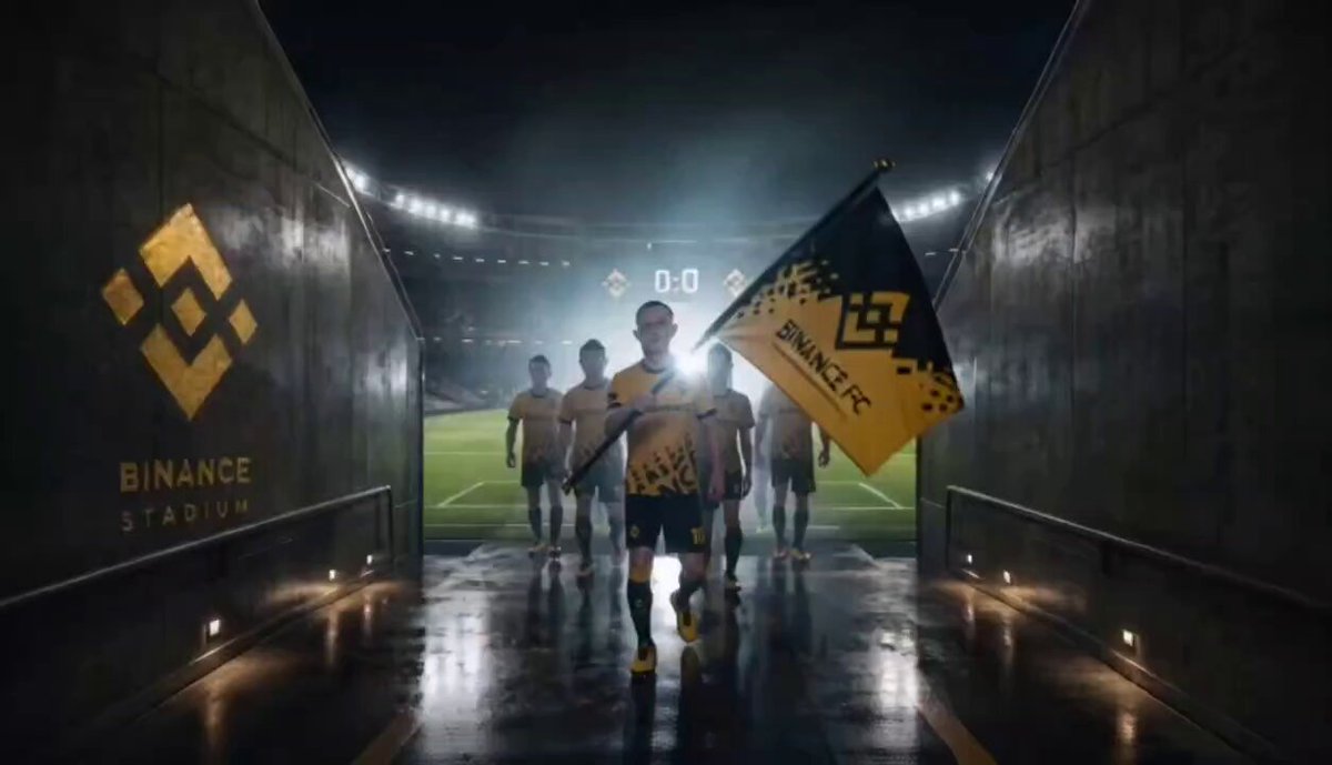 The image features a group of soccer players walking down an entrance to a stadium, with some of them holding a flag. The players are wearing yellow jerseys, which stand out against the dark background. In the foreground, there's a sign that reads "Binance Stadium", indicating the location or event taking place in this area.