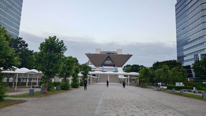 A wide plaza leading to a distinctive modern building with an angular, winged roof structure, flanked by trees and tall glass skyscrapers under a cloudy sky.
