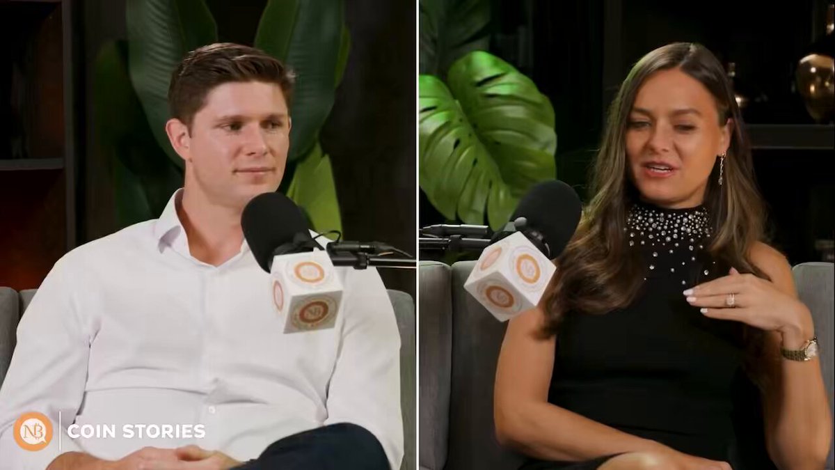 In this image, two people are sitting down while being interviewed or participating in a video recording session that was likely done by the host of the channel, "NZL Coin Stories". The two main characters in the image appear to be male and female, with one person holding onto a microphone and the other looking into a camera. There is also an umbrella visible in the scene, which might suggest that they are indoors or outdoors where it has been raining.
