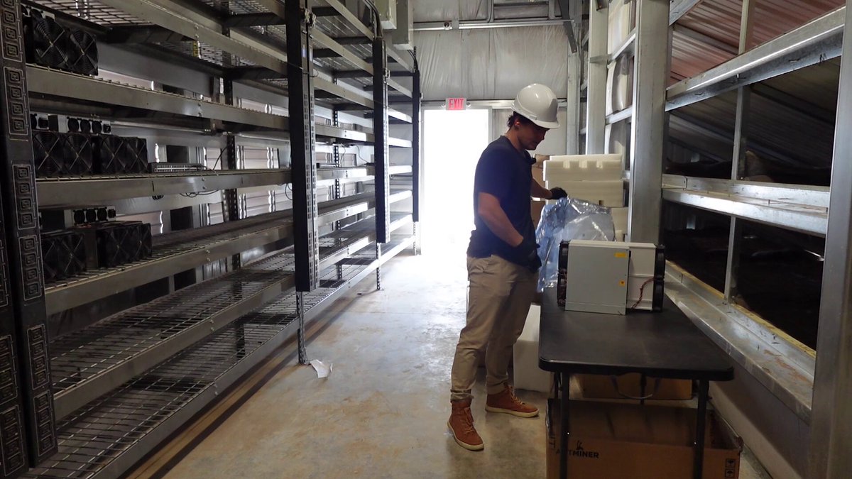 The image shows a man wearing a hard hat, standing next to a table with a computer monitor or laptop on top of it. He appears to be working at a desk within an industrial setting, possibly in a warehouse or factory environment. The scene also includes several shelves filled with various items such as boxes and other objects, suggesting that the area is used for storage purposes.