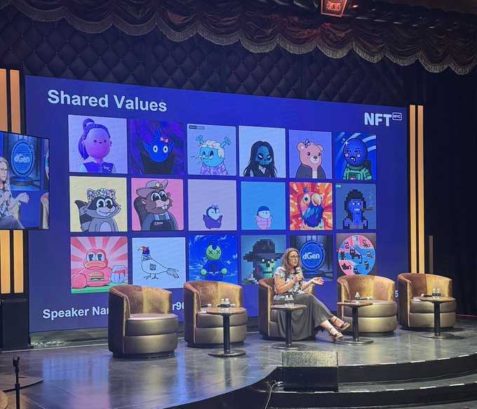 A stage presentation with a large screen displaying 'Shared Values' and 'NFT NYC', featuring a grid of various cartoon characters alongside a speaker seated in a leather chair.